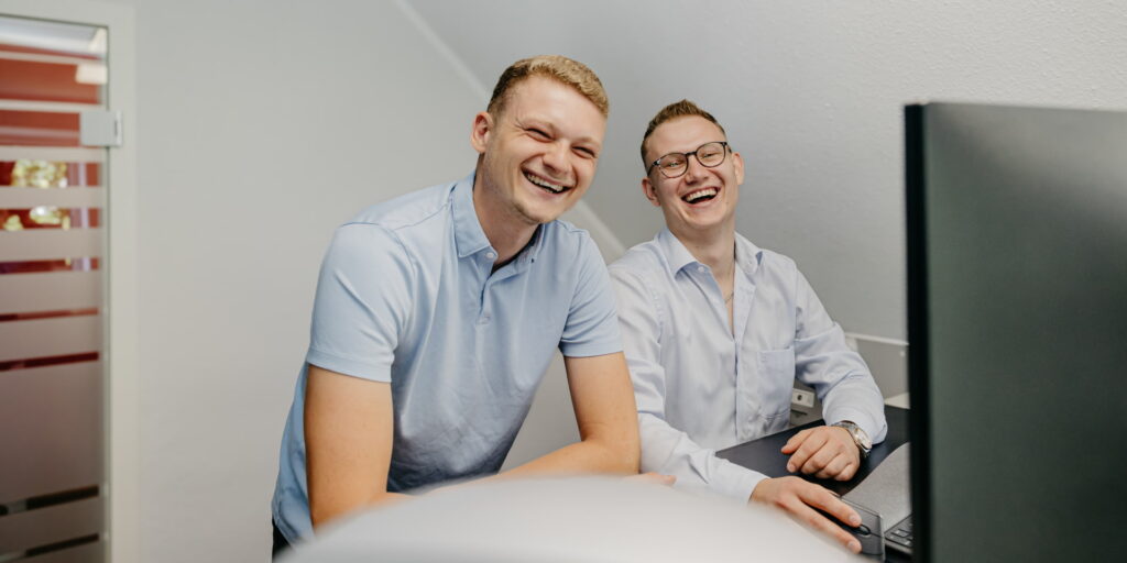 Zwei Kollegen stehen gemeinsam vor einem Bildschirm und lachen in die Kamera.