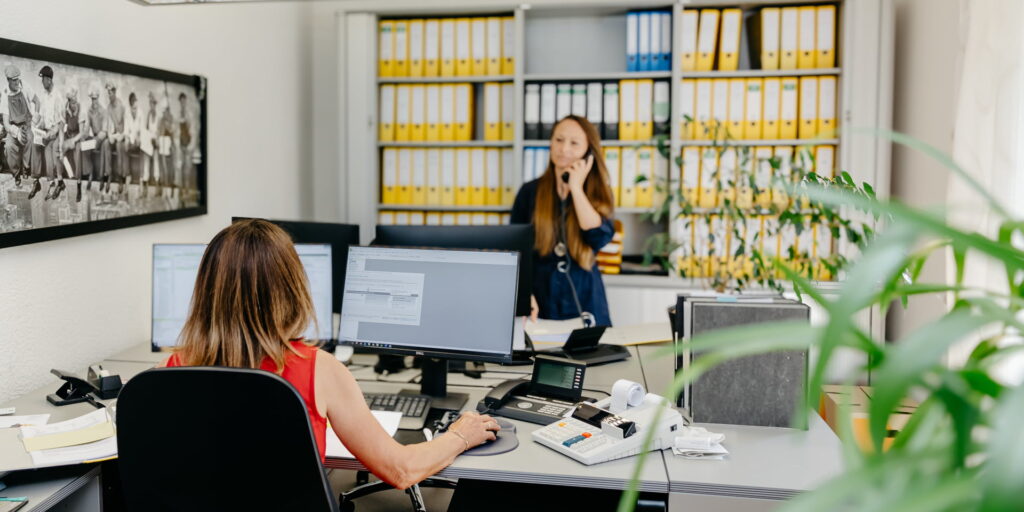 Eine Mitarbeiterin sitzt mit dem Rücken zur Kamera an ihrem Schreibtisch und arbeitet an ihrem PC. Eine Kollegin steht ihr gegenüber und telefoniert.