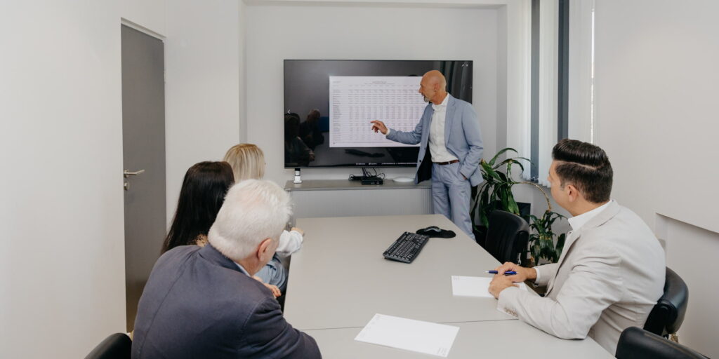 Ein Mitarbeiter zeigt auf einen großen Bildschirm in einem Besprchungsraum und 4 Kollegen hören und sehen ihm zu.