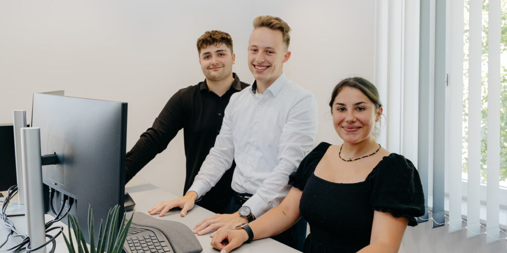 Drei Auszubildende stehen gemeinsam an einem Schreibtisch und lächeln in die Kamera.