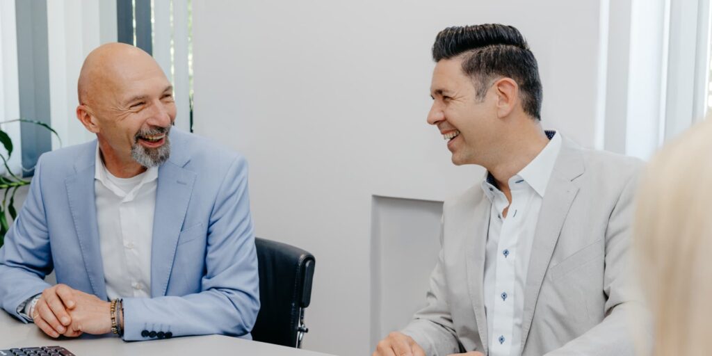 Zwei Kollegen sitzen gemeinsam an einem Tisch und lachen sich an.