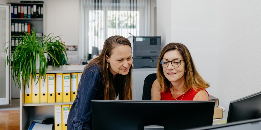 Zwei Kolleginnen betrachten gemeinsam einen Bildschirm und lächeln.