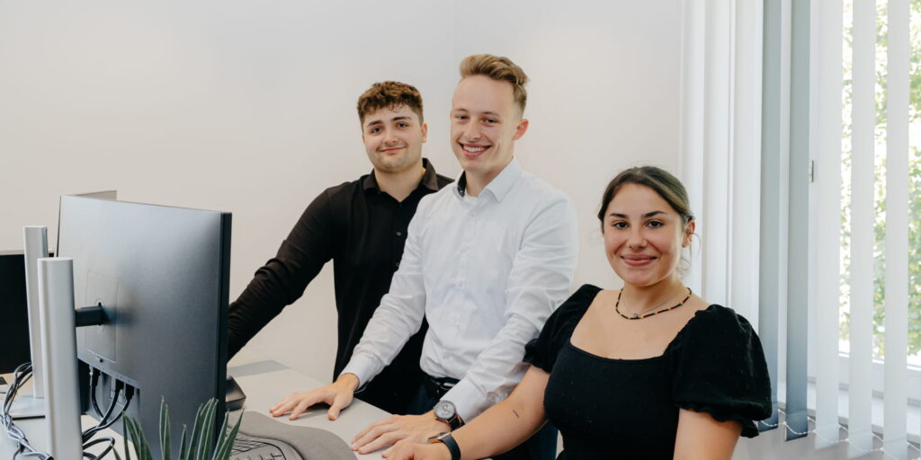 Drei Auszubildende stehen gemeinsam an einem Schreibtisch und lächeln in die Kamera.